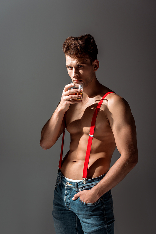 shirtless man holding glass of whiskey and standing with hand in pocket isolated on grey