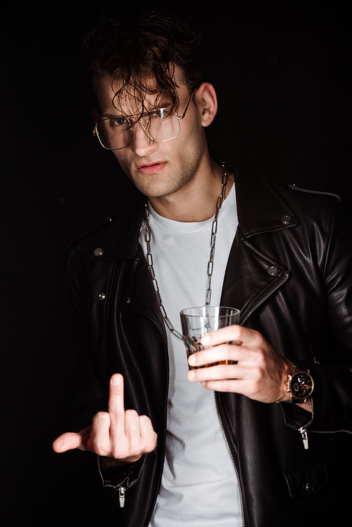 selective focus of stylish man in eye glasses holding whiskey and showing middle finger isolated on black