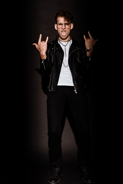 trendy man in glasses showing rock sign while sticking out tongue on black