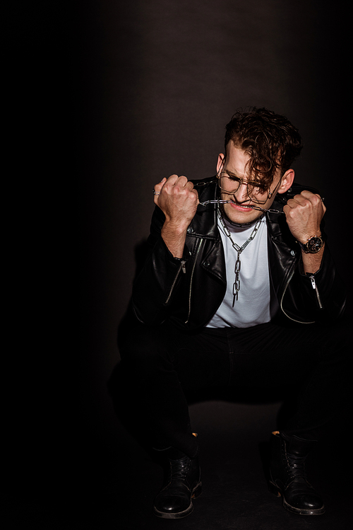 emotional man in glasses and leather jacket biting chain on black