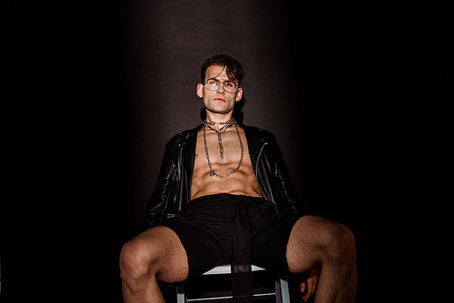 sexy and brutal young man in glasses and leather jacket sitting on black