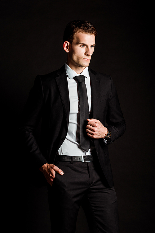 handsome businessman in formal wear standing with hand in pocket isolated on black