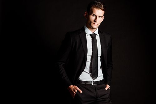 handsome businessman in formal wear standing with hands in pockets isolated on black