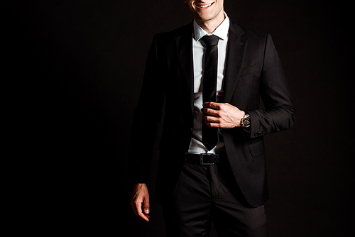 cropped view of happy businessman touching tie isolated on black