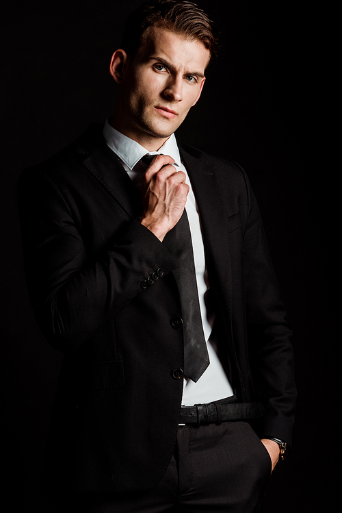 handsome businessman man touching tie while standing with hand in pocket isolated on black