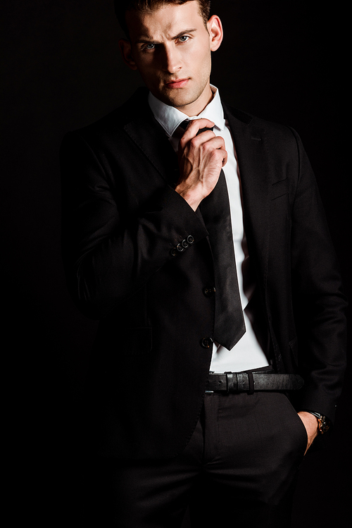 handsome businessman touching tie and standing with hand in pocket isolated on black