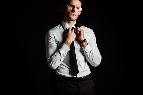 handsome man in glasses touching tie isolated on black