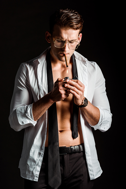 handsome man in glasses holding lighter near cigar and smoking isolated on black