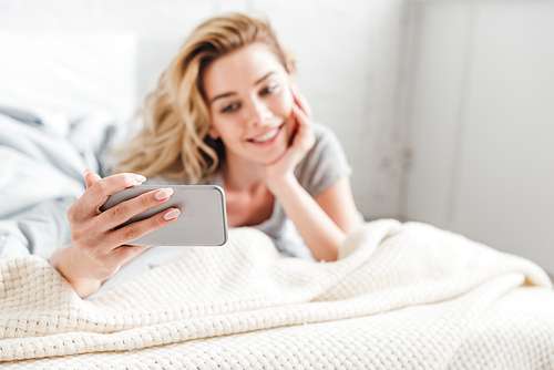 selective focus of happy girl taking selfie on smartphone while lying on bed