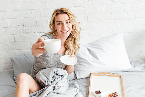 selective focus of happy woman holding cup of coffee and saucer in bed