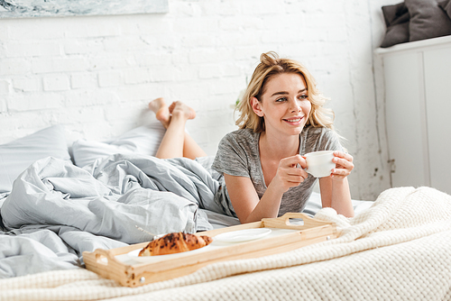 selective focus of happy girl holding cup of coffee near tray with tasty croissant on bed