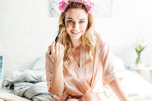 cheerful girl in hair curlers holding cosmetic brush near face