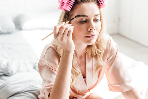 beautiful woman in pink hair curlers styling eyebrows in bedroom