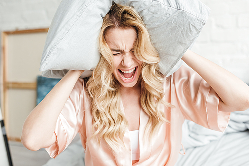 emotional woman in silk robe holding pillow screaming in bedroom