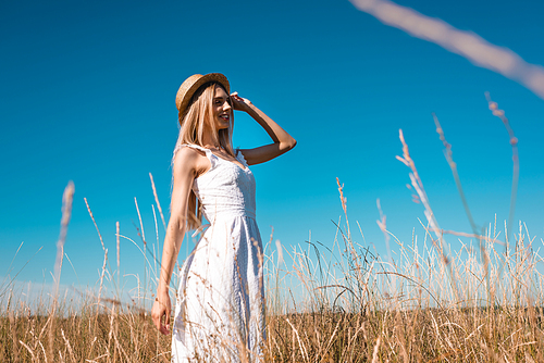 selective focus of stylish young woman in white dress touching straw hat and looking away against blue sky