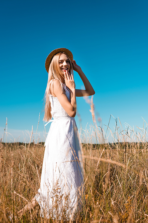 selective focus of sensual woman in white dress touching straw hat and holding hand near chin while
