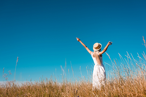 back view of woman in white dress and straw hat standing with outstretched hands in grassy field against blue sky