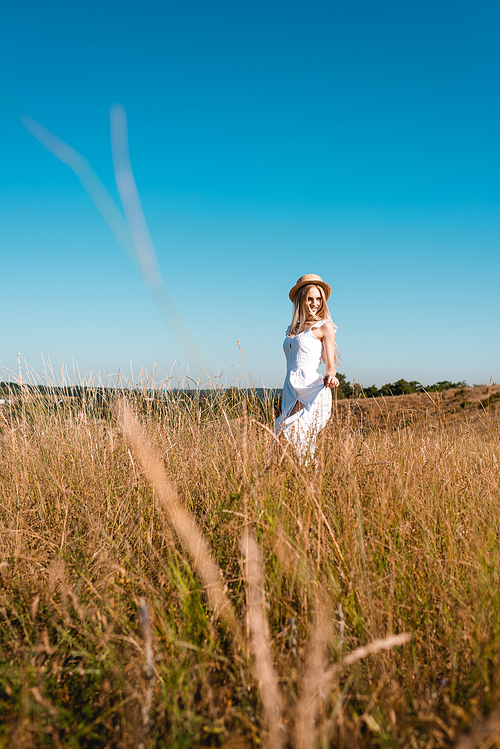 selective focus of stylish woman in white dress and straw hat standing in field against blue sky