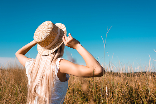 back view of blonde woman in summer outfit touching straw hat in grassy meadow against blue sky, selective focus