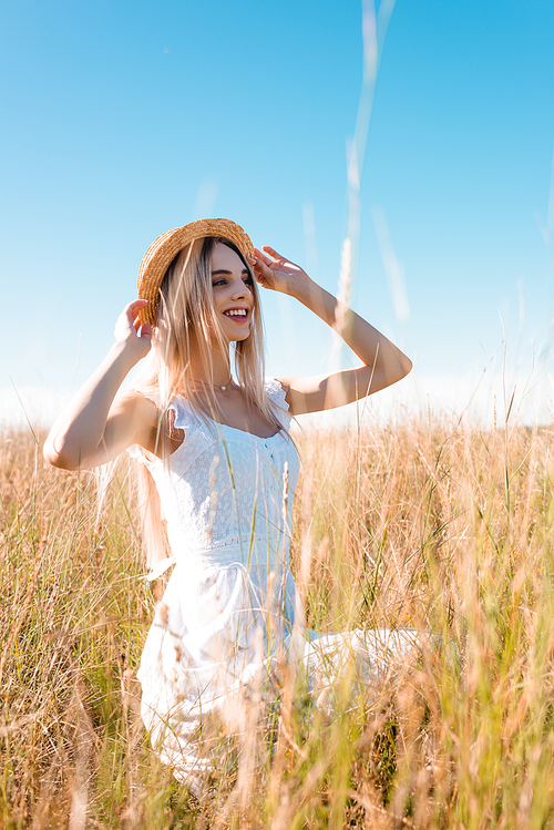 selective focus of stylish blonde woman in white dress looking away and touching straw hat while sitting in grassy meadow