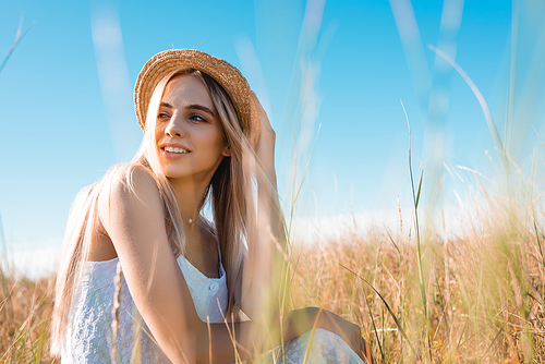 selective focus of stylish blonde woman looking away while sitting in field and touching straw hat