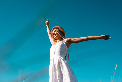 low angle view of woman in white dress and straw hat looking away while standing against blue sky with outstretched hands