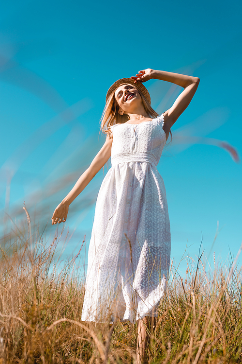selective focus of woman in white dress touching straw hat while standing with outstretched hand against blue sky