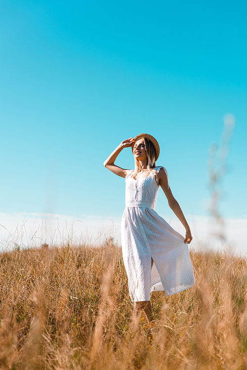 selective focus of stylish woman touching white dress and straw hat while standing in field with closed eyes