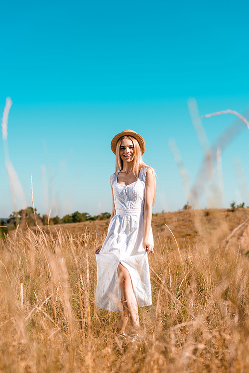 selective focus of sensual blonde woman in white dress and straw hat  while posing in grassland