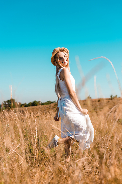 selective focus of woman in white dress and straw hat standing on one leg while posing in meadow