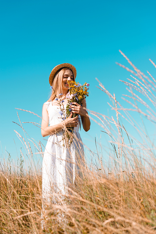 selective focus of blonde woman in straw hat and white dress holding bouquet of wildflowers against blue sky