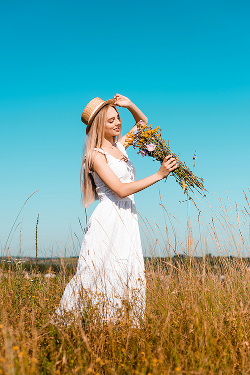 selective focus of sensual woman in white dress touching straw hat while holding wildflowers against blue sky