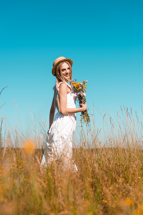 selective focus of sensual woman in white dress and straw hat holding wildflowers and  in grassy field