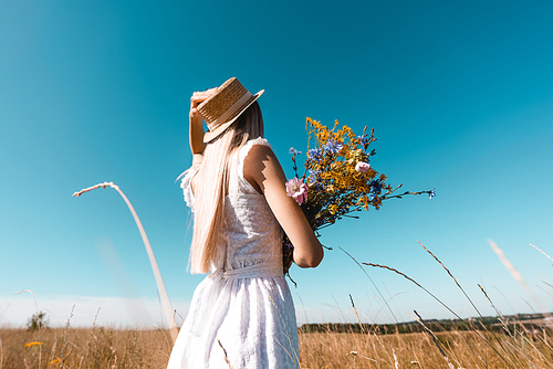 selective focus of young woman in white dress touching straw hat while holding bouquet of wildflowers against blue sky