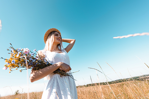 high angle view of young woman with wildflowers touching straw hat while looking away against blue sky