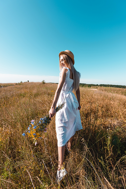 young woman in white dress and straw hat walking in grassy meadow with bouquet of wildflowers