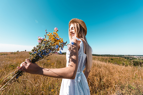 blonde woman in straw hat and white dress holding bouquet of wildflowers in meadow against blue sky