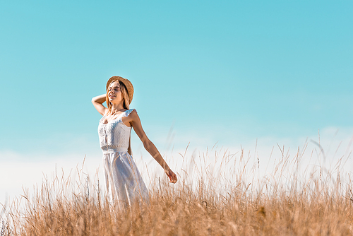 selective focus of sensual woman in white dress touching straw hat while standing with outstretched hands against blue sky