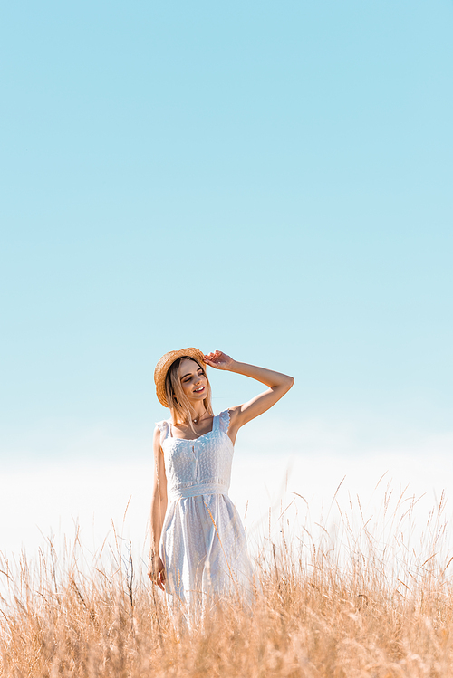 young woman in white dress standing on grassy hill, touching straw hat and looking away against blue sky