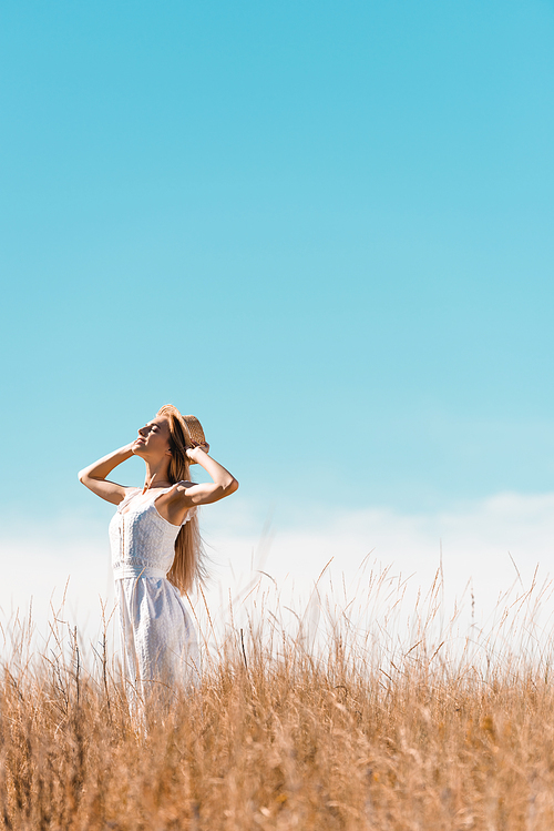 selective focus of stylish woman in white dress touching straw hat while standing with raised head against bule sky