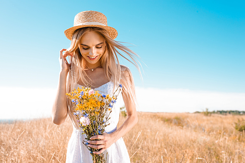 sensual young woman in white dress and straw hat holding wildflowers and touching hair in grassy meadow