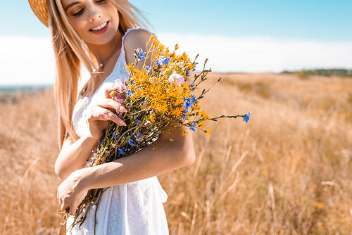 selective focus of young blonde woman holding bouquet of wildflowers in meadow