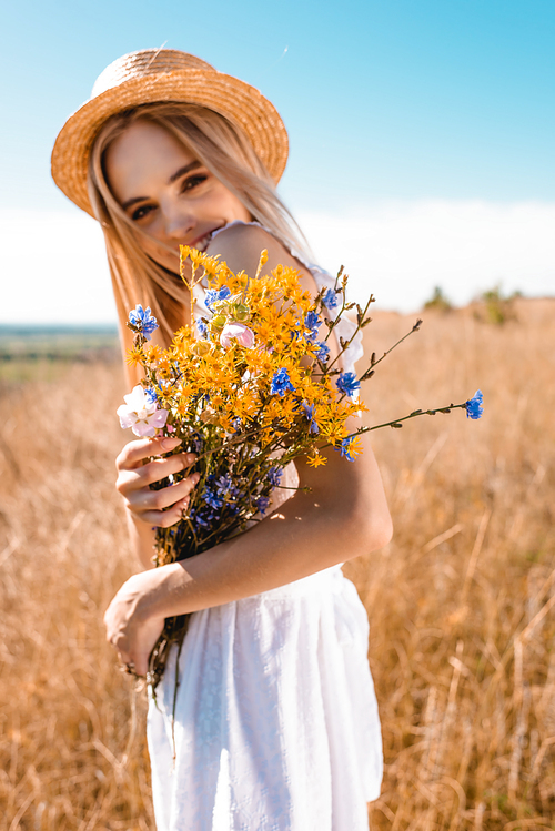 selective focus of blonde woman in straw hat holding wildflowers while  in grassy meadow