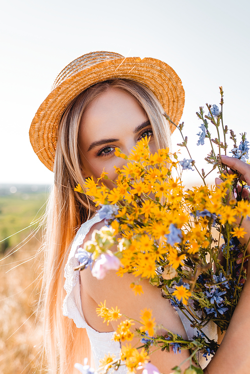 sensual blonde woman in straw hat holding wildflowers and  against sky