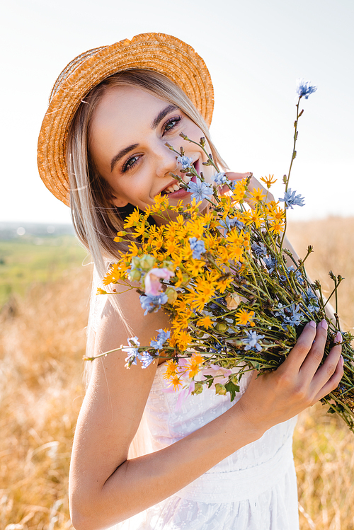 young blonde woman in white dress and straw hat holding wildflowers and  in grassy field