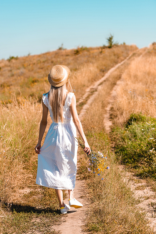 back view of young woman in white dress and straw hat holding wildflowers while walking on road in meadow