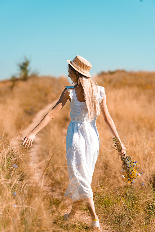 back view of young woman in white dress and straw hat holding wildflowers while walking on road in meadow