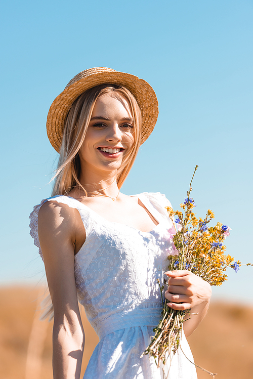 young woman in white dress and straw hat holding bouquet of wildflowers and  against blue sky
