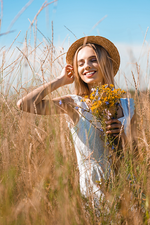 selective focus of young sensual woman touching straw hat while holding wildflowers in grassy field