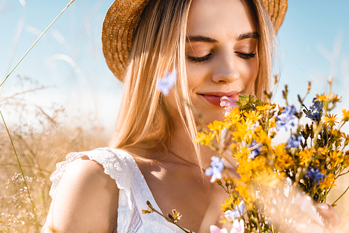 selective focus of sensual woman in straw hat smelling bouquet of wildflowers with closed eyes